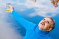 An elderly man clearly enjoys beer near the water with reflected clouds in autumn