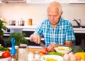 Elderly man cuts vegetables for salad at the table in the kitchen Royalty Free Stock Photo