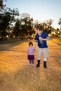 An Elderly Man with Downs Syndrome Holds the Hand of a Young Girl as they Walk Through a Park While the Sun Sets Behind Them