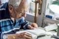 Elderly man with glasses reading writings in notebook near the window at home