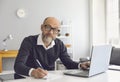 An elderly man with glasses is working he is studying he has a laptop he writes notes in a notebook while sitting at a Royalty Free Stock Photo