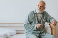 Elderly with gray beard and hair sits in pajamas on the bed with glasses in hand