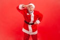 Elderly man with gray beard in santa claus costume keeping palm over head and looking attentively