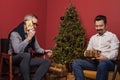 Elderly man and his son smiling and holding Christmas presents gold boxes with ribbons on red wall background and Christmas tree