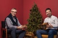 Elderly man and his son smiling and showing Christmas presents gold boxes on red background and Christmas tree