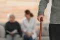 Elderly man holding walking cane and blurred caregiver with senior woman on background, focus on hand.