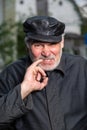 An elderly man in outdated black clothes and a leather black cap is picking his little finger in his tooth. Portrait