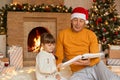 Elderly man reading book with his granddaughter in living room decorated for Christmas, family sitting on floor near fireplace and Royalty Free Stock Photo