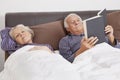 Elderly man reading book while spouse sleeping besides in bedroom