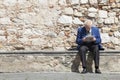 Elderly man reading and sitting on a stone bench. Stones wall