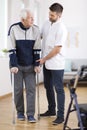 Elderly man walking on crutches and a helpful male nurse supporting him