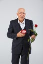 An elderly man on a white background, in the studio, in a black smart suit with a rose in his hand and a red gift box. Royalty Free Stock Photo