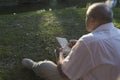 An elderly man in a white shirt is sitting on a blanket, on the ground in a park and reading an interesting book. A pensioner Royalty Free Stock Photo