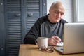 Elderly man working on laptop, looking at screen, makes notes in a notebook and drinking coffee Royalty Free Stock Photo