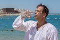 An elderly man 45-50 years old drinks water from a plastic bottle against the background of the sea. Concept: thirst and dehydrati