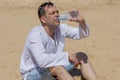 An elderly man 45-50 years old greedily drinks water from a plastic bottle sitting on the sand in the desert. Concept: thirst and