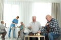 Elderly men playing chess while nurse serving breakfast to women. Assisting senior people