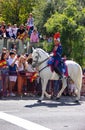 An elderly military man in a red and blue uniform rides a white horse past the citizens of Granada