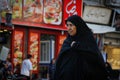 An elderly Muslim Iranian woman covered in a black veil chador goes along the city street among the shops
