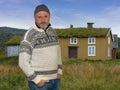 A Norwegian with a sweater is standing in a meadow in front of a typical old wooden house with a grass roof. Royalty Free Stock Photo