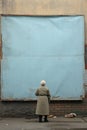 An elderly old woman, seen from behind, gazes at an empty and weathered blue poster, hinting at stories of the past