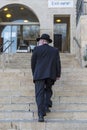 An elderly Orthodox Jew in a black suit and hat walks up the stairs in Jerusalem. Vertical. Back view