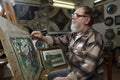 Elderly painter with beard and glasses draws a flowers picture by oil paint in art workshop