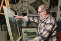 Elderly painter with beard and glasses draws a flowers picture by oil paint in art workshop