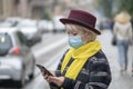 An elderly pensioner 60-65 years old in a medical mask dials a number on the phone against the backdrop of the city landscape. The