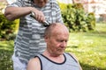 Woman is doing haircut with hair clipper to senior man outdoors at garden