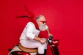 Elderly santa man moving fast to party by vintage moped wear white jumper and trousers red background