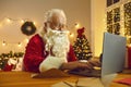 Elderly Santa with white beard in red costume chatting and congratularing online on laptop