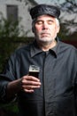 An elderly senior gray-haired man in old black clothes and a black cap holds a glass of beer and enjoys the taste of Royalty Free Stock Photo