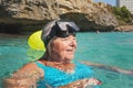 Elderly senior woman smiling in calm calm sea on sunny day, fogged diving mask on head, rocky cliff background