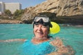 Elderly senior woman smiling in calm calm sea on sunny day, fogged diving mask on head, rocky cliff background