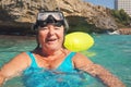 Elderly senior woman smiling in calm calm sea on sunny day, fogged diving mask on head, rocky cliff background