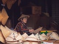 An elderly shop keeper surrounded by a heap of oriental hats.