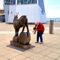 Elderly white-haired lady with headscarf and red jacket takes photo of wooden sculpture of chamois on Austrian platform of