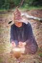 An elderly witch in a hat in the woods, sitting on the ground, brewing a witch`s potion with fly agarics Royalty Free Stock Photo
