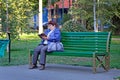 An elderly woman in a blue jacket is reading a book on a bench.