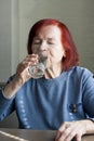 elderly woman drinking mineral water from glass. Portrait of red-haired senior woman in blue jumper, quenches thirst, sitting at Royalty Free Stock Photo