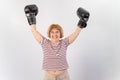 An elderly woman in fighting gloves raises her hands up on a white background. Victory in a duel