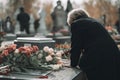 An elderly woman with gray hair leans over a granite tombstone and puts a bouquet of roses on it. Mourning for a loved one