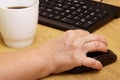 An elderly woman hand on a computer mouse. The old grandmother works behind the computer keyboard and drinks coffee Royalty Free Stock Photo