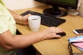 An elderly woman hand on a computer mouse. The old grandmother works behind the computer keyboard and drinks coffee Royalty Free Stock Photo