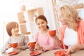 Elderly woman and happy grandson and granddaughter drink tea from red mugs in kitchen.