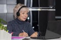 An elderly woman in her 60s and 65s is sitting in front of her laptop with headphones on. Concept: remote work, learning foreign l Royalty Free Stock Photo