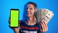 Elderly woman holding phone with green screen in one and money in another hand. Lottery win