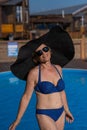 An elderly woman in a huge straw hat and sunglasses is sunbathing by the pool. Retiree on vacation.