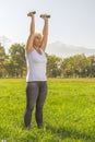 Elderly woman lifts dumbbells while doing fitness in a city park against the backdrop of mountains